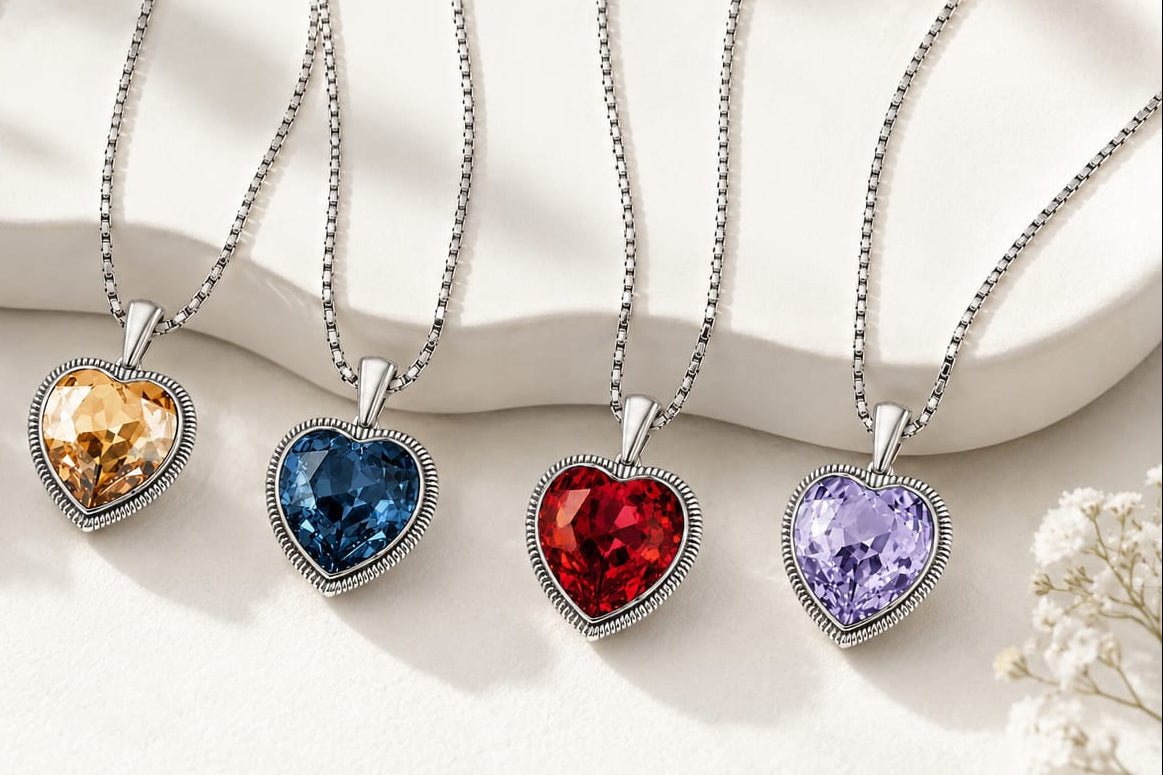 Four heart-shaped pendant necklaces with silver-toned chains and bezel settings, each holding a different colored gemstone (orange, denim blue, red ruby, and lavender), arranged on a white ceramic surface with soft floral accents.