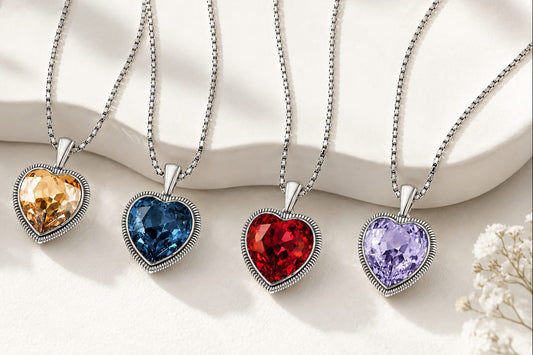 Four heart-shaped pendant necklaces with silver-toned chains and bezel settings, each holding a different colored gemstone (orange, denim blue, red ruby, and lavender), arranged on a white ceramic surface with soft floral accents.