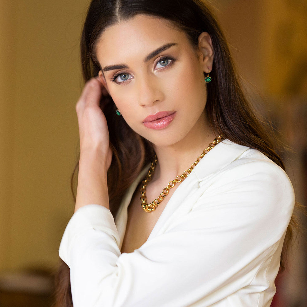 Woman wearing a gold necklace and emerald 18ct gold-plated earrings indoors.