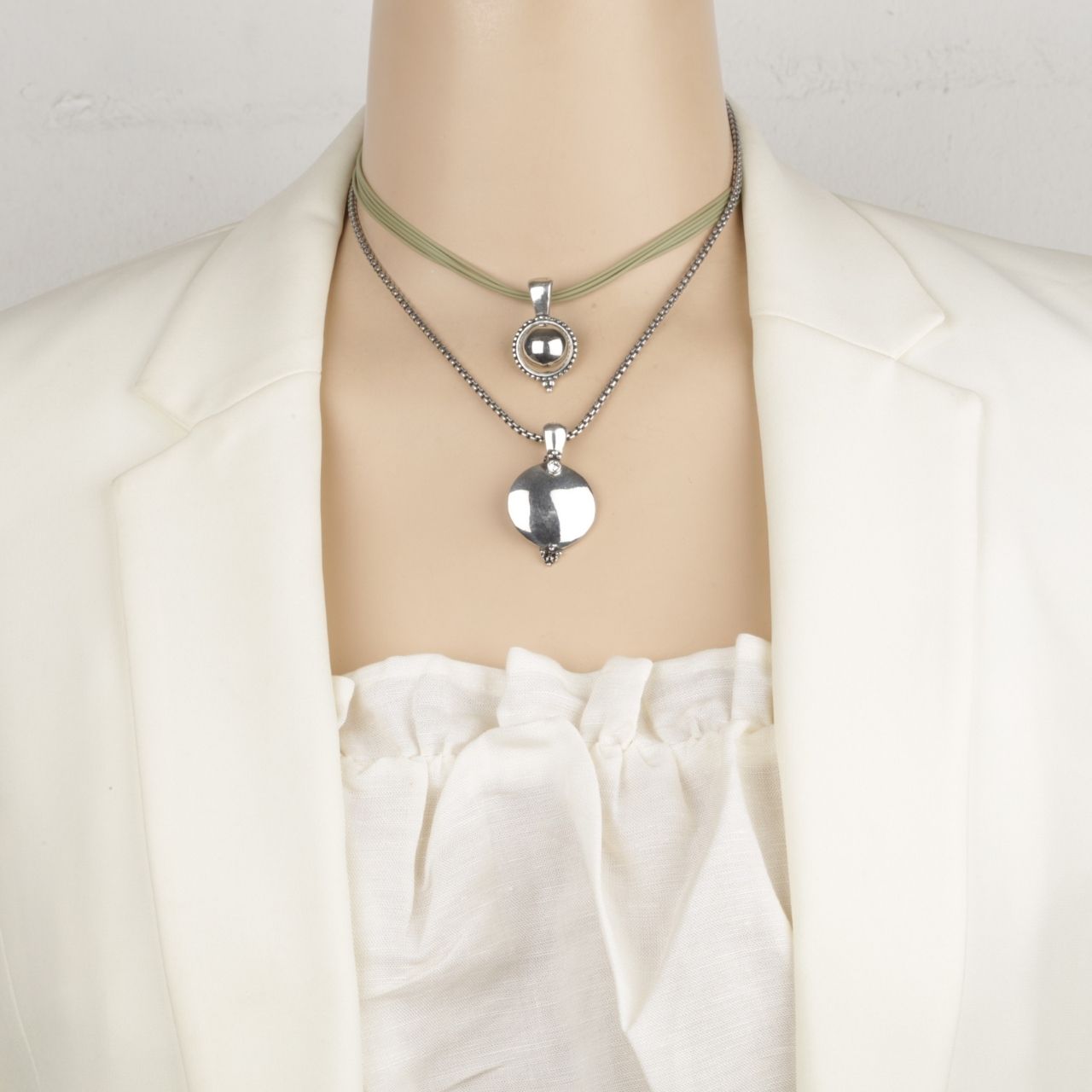 Mannequin wearing a silver necklace with a heart pendant and green leather necklace with a circular pendant against a white background