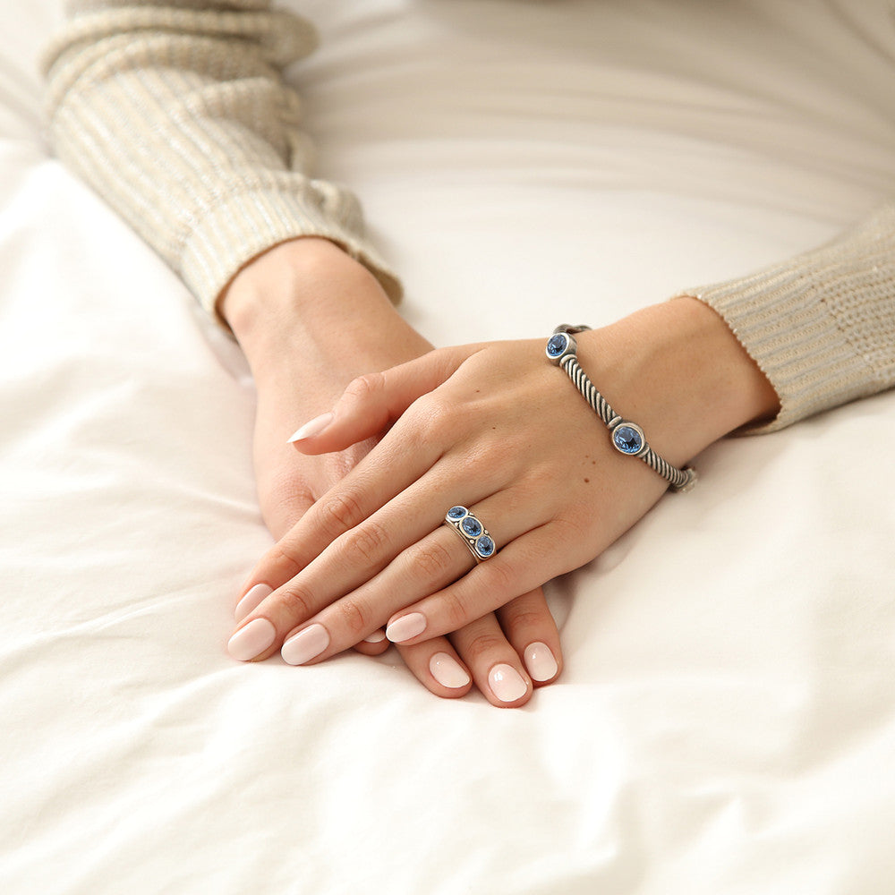 A close-up of a person's hands resting on white bedding, wearing a silver-toned twisted blue bracelet and matching ring with 3 oval blue stones.